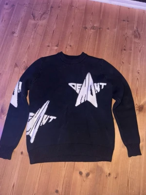 Demont tröja - Cool svart stickad tröja med stora vita grafiska tryck och texten 'DEMONT' på bröstet, ärmen och sidan. Tröjan har rund halsringning och ribbade muddar vid ärmslut och nederkant. Perfekt för dig som gillar streetwear och unik design.