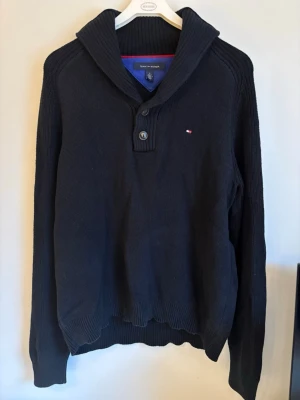 Mörkblå stickad tröja Tommy Hilfiger - Mörkblå stickad tröja från Tommy Hilfiger med hög krage och två knappar vid halsen. Liten broderad logga på bröstet. Tröjan har långa ärmar och ribbade muddar vid ärmslut och nederkant. Perfekt för kyliga dagar och snygg till jeans.