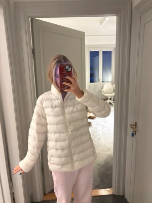 Vit pufferjacka med dragkedja - Säljer en stilren vit pufferjacka med hög krage och dragkedja framtill. Jackan har en quiltad design med horisontella sömmar och två sidofickor. Perfekt för kalla vinterdagar och ger en clean look.