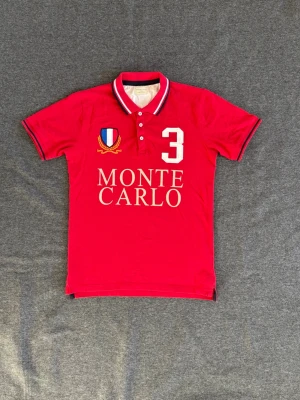 Röd Monte Carlo pikétröja - Snygg röd pikétröja från Monte Carlo med vit text och nummerdetalj framtill. Tröjan har klassisk krage med knappar och kontrastfärgade ränder på krage och ärmslut. Broderad sköld på bröstet och stor siffra 3 ger en sportig vibe.