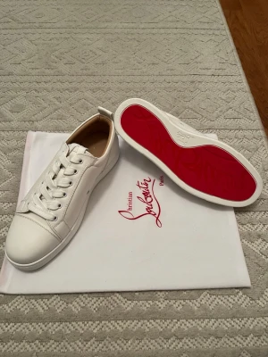 Vita Christian Louboutin - Säljer ett par vita sneakers från Christian Louboutin i skinn med ikonisk röd sula. Klassisk låg modell med rund tå, vita snören och diskreta detaljer. Perfekta för dig som vill ha en clean look med lyxig touch och den där signaturröda sulan som sticker ut.