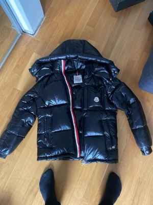 Svart Moncler dunjacka med huva - Pris kan diskuteras! Snygg svart dunjacka från Moncler med glansig finish, stor huva och klassisk logotyp på bröstet. Jackan har en dragkedja med röd, vit och blå detalj samt flera praktiska fickor. Perfekt för kalla vinterdagar och riktigt trendig stil.