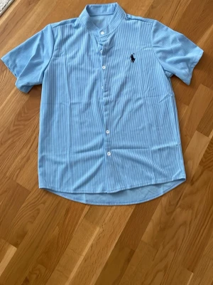 Ljusblå randig skjorta Polo Ralph Lauren - Kortärmad ljusblå Polo Ralph Lauren  skjorta med tunna vita ränder och klassisk knappstängning. Skjortan har en broderad logga på bröstet och en rak passform. Perfekt för en avslappnad men stilren look.