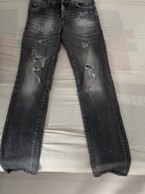 Svarta slitna jeans med distressed detaljer - Säljer ett par svarta jeans med coola slitningar och distressed detaljer framtill. Jeansen har en tvättad look med ljusare partier och raka ben. Klassisk femficksmodell och knappgylf. Perfekta för en avslappnad streetstil.