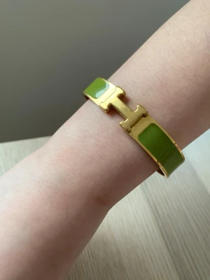Grönt  Clic H armband - Snyggt armband från  i modellen Clic H. Armbandet har guldfärgad metall och gröna emaljdetaljer. Den ikoniska H-detaljen framtill ger en exklusiv känsla och armbandet har en stilren, modern design som sticker ut.