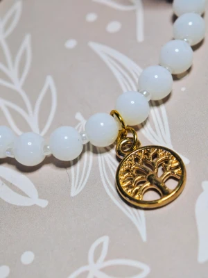 Vitt pärlarmband med guldfärgad berlock - Stilrent armband med vita runda glaspärlor och en guldfärgad berlock i form av ett livsträd. Armbandet är elastiskt och har en längd på 18 cm. Perfekt accessoar för att lyfta din outfit med en elegant touch.