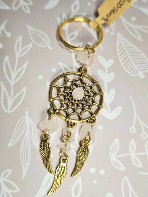 Drömfångare nyckelring i guld och rosenkvarts  - Säljer en snygg nyckelring formad som en drömfångare i guldfärgad metall med detaljer av ljusrosa stenar och tre hängande vingar. Perfekt accessoar för att piffa upp väskan eller nyckelknippan.