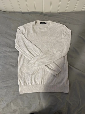 Dressman stickad tröja - En stilren ljusgrå tröja från Dressman med rund halsringning och långa ärmar. Tillverkad i mjukt och tunt material, perfekt för lager på lager. Tröjan har ribbade muddar vid ärmslut och nederkant för en clean look.