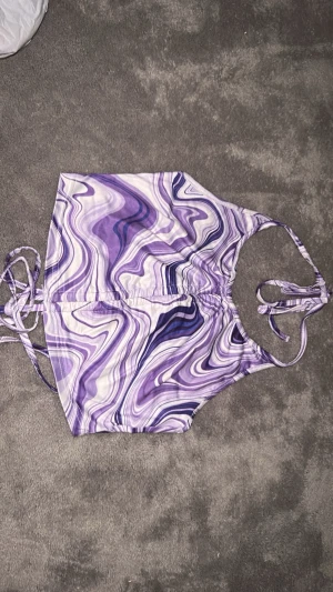 Lila swirl bikini med knytband - Säljer en bikini med swirlmönster i olika lila toner och vitt. Bikinin har smala knytband i sidorna och vid axlarna för justerbar passform. Materialet känns mjukt och stretchigt, perfekt för sommardagar på stranden.