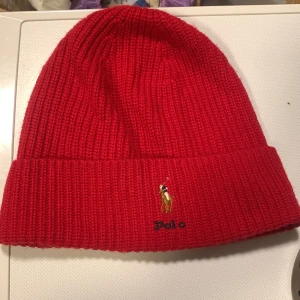 Röd ribbstickad mössa från Polo Ralph Lauren - Säljer en snygg röd ribbstickad mössa från Polo Ralph Lauren med uppvikt kant och den klassiska broderade loggan framtill. Perfekt för att hålla värmen och ge din outfit en färgklick. Materialet känns mjukt och stretchigt.
