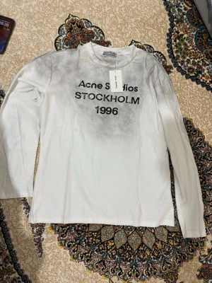 Vit långärmad topp Acne Studios - Vit långärmad topp från Acne Studios med svart text 'Acne Studios STOCKHOLM 1996' framtill. Tunn och mjuk i materialet, rund halsringning och diskret grå toning upptill. Perfekt för en trendig och avslappnad stil.