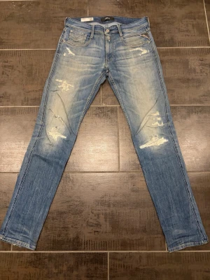 Blå slitna jeans från Replay - Säljer ett par blå jeans från Replay med snygga slitningar och tvättade detaljer. Modellen är smal med klassisk femficksdesign och dragkedjegylf. Perfekta för dig som gillar en avslappnad och trendig stil. Storlek 31/34
