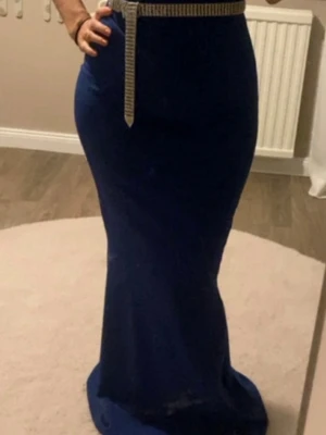 Marinblå långklänning - Snygg marinblå långklänning med figurnära passform och ett glittrigt bälte i midjan som ger extra detalj (men bältet medföljer inte med, man kan lägga till liknande bälte själv om man har hemma). Klänningen har ärmlös design och markerad midja, perfekt för dig som vill sticka ut lite extra.