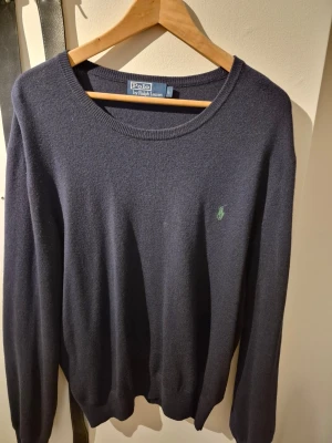 Mörkblå tröja från Polo Ralph Lauren - Klassisk mörkblå tröja från Polo Ralph Lauren med rund halsringning och diskret grön logga broderad på bröstet. Tröjan har långa ärmar och ribbade muddar vid ärmslut och nederkant. Perfekt för en stilren och enkel look.