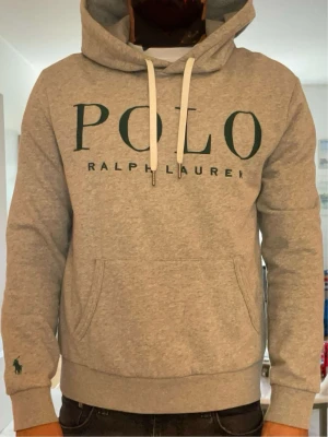 Ralph Lauren Hoodie - Snygg grå hoodie från Polo Ralph Lauren med stor logga i grönt framtill och klassisk känguruficka. Huva med vita dragsnören och liten grön broderad logga på ärmen. Tillverkad i mjuk bomullsblandning för maximal komfort.