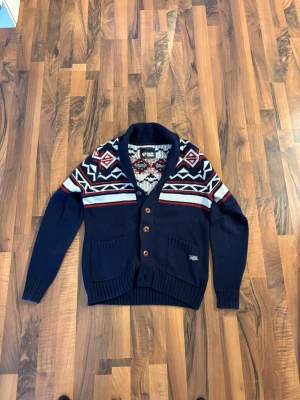 Mörkblå cardigan/kofta  med knappar  - Snygg mörkblå kofta med sjalkrage och knappar framtill. Koftan har ett klassiskt stickat mönster i vitt och rött över axlarna samt två fickor fram. Perfekt för dig som gillar retrovibbar och vill ha något både varmt och stilrent.