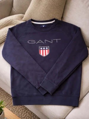Mörkblå sweatshirt från GANT - Snygg mörkblå sweatshirt från GANT med broderad logga och ett sköldmärke med röda och vita ränder samt tre vita stjärnor på bröstet. Tröjan har rund halsringning och långa ärmar. Perfekt för en avslappnad och stilren look. Storlek: S 160
