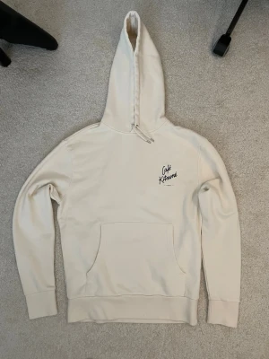 Beige hoodie från Café Kitsuné - Stilren beige hoodie från Café Kitsuné med broderad logga på bröstet. Klassisk känguruficka framtill och justerbar huva med dragsko. Perfekt för en avslappnad och trendig look.