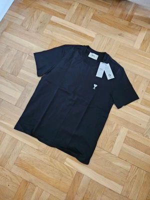 Svart t-shirt från Ami Paris - Svart t-shirt från Ami Paris med korta ärmar och en vit broderad logga med hjärta och bokstaven A på bröstet. Tillverkad i mjuk bomull och har en klassisk passform. Perfekt till jeans eller shorts.