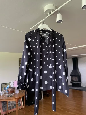 Svart prickig blus med knytband - Snygg svart blus med stora vita prickar och långa ärmar. Blusen har knytband framtill och en lös, avslappnad passform. Materialet ser ut att vara glansigt, troligtvis polyester eller satin. Perfekt för dig som gillar statement-plagg och mönster. Stl 36 - S 