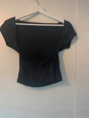 Topp med öppen rygg - Snygg svart  topp med öppen rygg och korta ärmar. Toppen har en figurnära passform och är tillverkad i ett mjukt, stretchigt material som sitter bekvämt på kroppen. Perfekt för dig som vill ha en enkel men stilren look. Storlek S