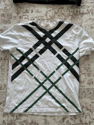 Vit t-shirt från burberry - Vit t-shirt från Anerkjendt med ett stort grafiskt mönster i svart och mörkgrönt över hela framsidan. Klassisk rund hals och korta ärmar. Perfekt för dig som gillar unika prints och modern stil.