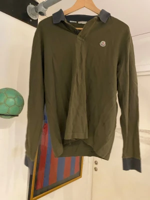Olivgrön Moncler långärmad skjorta - Snygg olivgrön långärmad skjorta från Moncler med mörkgrå krage och muddar. Slim fit-modell med klassisk knappslå och broderad Moncler-logga på bröstet. Perfekt för dig som gillar stilrena och exklusiva plagg.