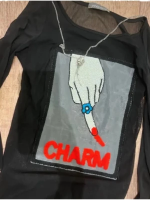 Svart mesh topp  - Cool svart mesh topp från Vince Camuto med lång ärm och stort tryck framtill av en hand med blå ring och röd texten 'CHARM'. Toppen har en unik design och är genomskinlig med en edgy vibe.