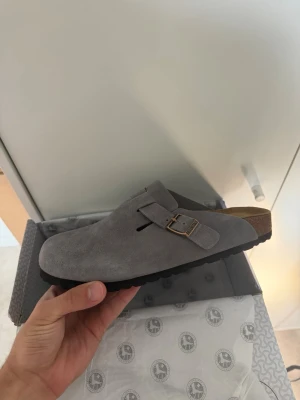 Grå mockatofflor från Birkenstock - Säljer ett par gråa tofflor från Birkenstock i mjuk mocka med justerbart spänne i metall. Klassisk slip-in modell med korksula och svart yttersula. Perfekta för dig som gillar stilren och bekväm design.