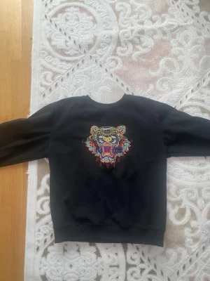 Kenzo tröja - Svart hoodie från Kenzo med ikonisk tigerbrodyr i flera färger på bröstet och texten 'KENZO PARIS' under. Klassisk passform med rund hals och ribbade muddar. Perfekt statement-plagg för dig som gillar streetwear och vill sticka ut.