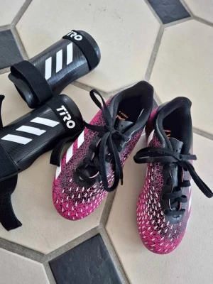 Adidas rosa och svarta fotbollsskor - Säljer ett par snygga Adidas fotbollsskor i rosa och svart med vita detaljer. Skorna har snörning och en cool struktur på ovandelen för bättre bollkontroll. Perfekta för dig som vill sticka ut på planen! Skorna är i syntetmaterial och har klassisk Adidas-design.