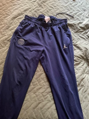 Mörkblå PSG x Jordan träningsbyxor - Snygga mörkblå träningsbyxor från Jordan i samarbete med PSG. Byxorna har resår i midjan med dragsko, PSG-logga på ena benet och Jumpman-logga på andra. Tillverkade i ett lätt och bekvämt material, perfekta för träning eller chill. Storlek L