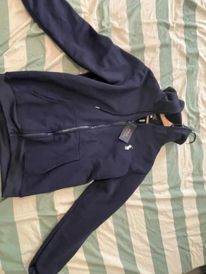Mörkblå Polo hoodie  - Snygg mörkblå hoodie från Polo Ralph Lauren med dragkedja framtill och klassisk vit logga på bröstet. Hoodien har huva med dragsko och känguruficka. Perfekt för en avslappnad och stilren look.