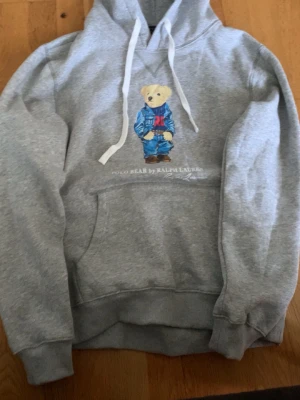 Grå hoodie Polo Bear Ralph Lauren - Grå hoodie från Ralph Lauren med Polo Bear-tryck framtill. Klassisk känguruficka, vit dragsko och ribbade muddar. Mjuk bomullsblandning och avslappnad passform. Perfekt för en chill och stilren look. Har bara haft på mig den en gång. Säljer pågrund av storlek 