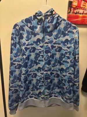 Blå camo hoodie från BAPE - Säljer en blå camo hoodie från BAPE med hel dragkedja och huva. Hoodien har ett unikt camomönster i olika blå nyanser och detaljer som apansikte och broderade bokstäver på huvan. Perfekt för dig som gillar streetwear och vill sticka ut.