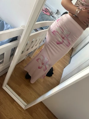 Ljusrosa långkjol med blommor - Supersnygg ljusrosa långkjol med stora blommiga tryck i vitt och rosa. Kjolen har en tight passform och är gjord i ett mjukt, stretchigt material som sitter skönt på kroppen. Perfekt för sommarens alla tillfällen!