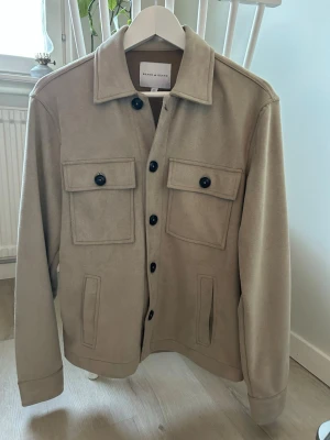 Beige overshirt från Brand Island  - Stilren beige overshirt med klassisk krage och svarta knappar från Sands & Brands. Två stora bröstfickor med lock och knappar samt två sidofickor. Tillverkad i ett mjukt material som ger en clean och modern look.