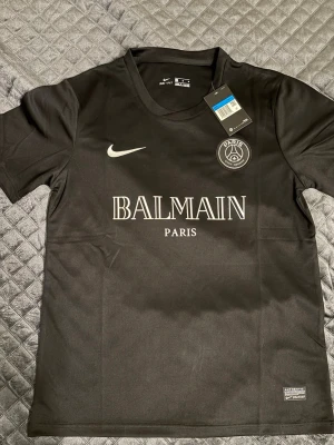 Svart PSG x Balmain tröja Nike - Snygg svart t-shirt från Nike i samarbete med PSG och Balmain. Tröjan har korta ärmar, rund hals och tryck med Balmain Paris och PSG-logga på bröstet. Tillverkad i ett lätt och bekvämt material, perfekt för sport eller streetwear.