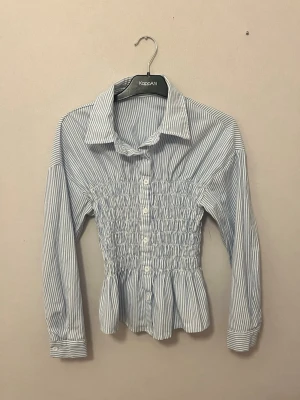 Blårandig smockad blus KappAhl - Snygg blus från KappAhl med blå och vita ränder, smockad midja och knappar framtill. Klassisk krage och långa ärmar med manschett. Tillverkad i ett lätt bomullsliknande material som ger en fräsch och trendig look.