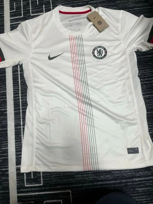 Vit Chelsea Nike fotbollströja - Chelsea FC matchtröja i vitt med tunna svarta och röda linjer längs framsidan. Tröjan har korta ärmar, rund hals och Chelsea-logga på bröstet. Tillverkad i Dri-FIT-material som andas, perfekt för träning eller match ingen namn på den. Pls buy