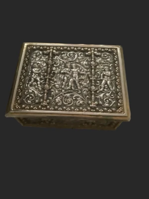 Dekorativt antikt smyckesskrin med reliefmönster - Säljer ett rektangulärt skrin med detaljerat reliefmönster på locket. Asken har antik känsla med ornament och figurer i silverfärg. Perfekt för småsaker eller som inredningsdetalj. Insidan är enkel och utan fack. Går att klä om i limmad textil och till exempel en skuren spegelbit som passar i botten för den händige fixaren. Eller bara behålla den äldre patinan som den är. 