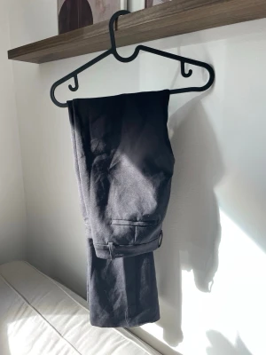 Svarta lågmidjade kostymbyxor BIKBOK  - Snygga svarta kostymbyxor från BIKBOK med utvidgade ben och lådmidjad. Byxorna har diskreta fickor bak och bälteshällor i midjan. Perfekta för en clean och stilren look. Materialet är mjukt och följsamt för extra komfort.