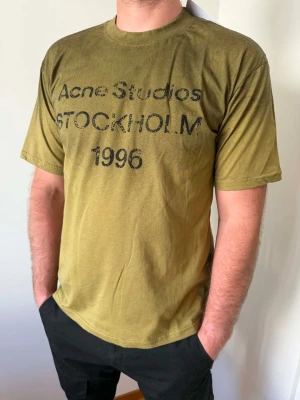 Olivgrön Acne Studios t-shirt - Säljer en olivgrön t-shirt från Acne Studios med tryck 'STOCKHOLM 1996' på bröstet. T-shirten har rund hals, korta ärmar och en relaxed passform. Tillverkad i mjuk bomull som känns skön mot huden. Perfekt för en avslappnad och trendig look.