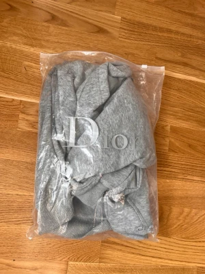 Grå hoodie  - Snygg grå hoodie med broderad logga på bröstet och klassisk känguruficka. Mjuk bomullsblandning och ribbade muddar vid ärmslut och midja. Perfekt för en chill och stilren look.