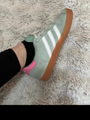 Adidas gazelle sneakers i mintgrönt och rosa - Snygga Adidas gazelle sneakers i mintgrönt mocka med vita ränder och detaljer i rosa på hälen. Skorna har klassisk gummisula i brunt och vita skosnören. Perfekt för dig som gillar färg och retrostil.