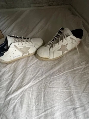 Golden Goose vita sneakers med stjärna - Snygga vita sneakers från Golden Goose med den ikoniska stjärnan i grå mocka på sidan och svart hälparti. Skorna har klassisk låg modell, vita skosnören och slitstark gummisula. Perfekta för en trendig och avslappnad look. Int super bra skick framre sulan sitter löst och är limmad men kan åka upp under frakten