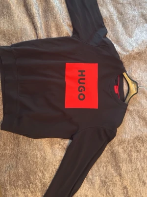 Svart sweatshirt från Hugo Boss - Cool svart sweatshirt från Hugo Boss med stor röd logga på bröstet. Tröjan har rund halsringning och långa ärmar. Tillverkad i mjukt material som känns skönt mot huden. Perfekt för dig som gillar stilrena och trendiga plagg.