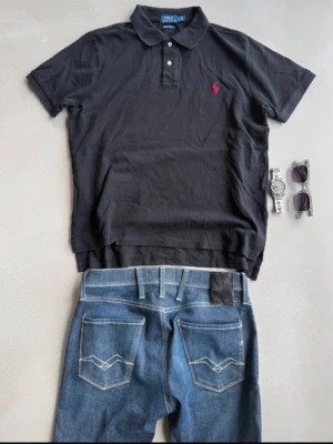 Svart pikétröja från Ralph Lauren - Klassisk svart pikétröja från Ralph Lauren med röd broderad logga på bröstet. Tröjan har krage, korta ärmar och knäppning med två knappar framtill. Perfekt för en stilren och avslappnad look. Står som size XL men sitter mer som en M, fråga gärna om mått vid behov :) 