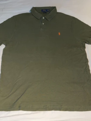 Olivgrön pikétröja från Ralph Lauren - Snygg olivgrön pikétröja från Ralph Lauren med klassisk krage och två knappar. Tröjan har den ikoniska orangea loggan broderad på bröstet. Tillverkad i bomull för en skön och avslappnad känsla.