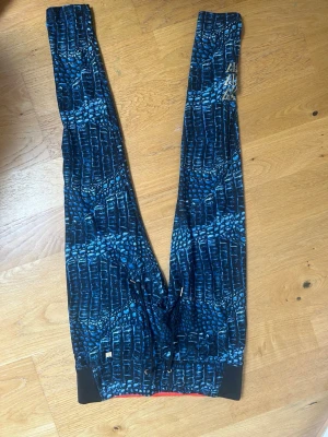 Blåmönstrade leggings från PowerWoman - Säljer ett par leggings från PowerWoman med blått och svart reptilmönster. De har dragsko i midjan, svarta detaljer vid benslut och en liten metallogo på sidan. Materialet är stretchigt och passar perfekt till träning eller chill.